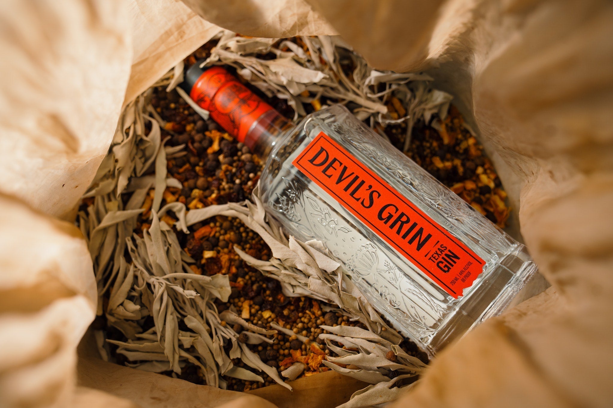 Devil's Grin Texas Gin bottle resting in a bag of dried botanicals, spices, and sage used in the gin distillation process