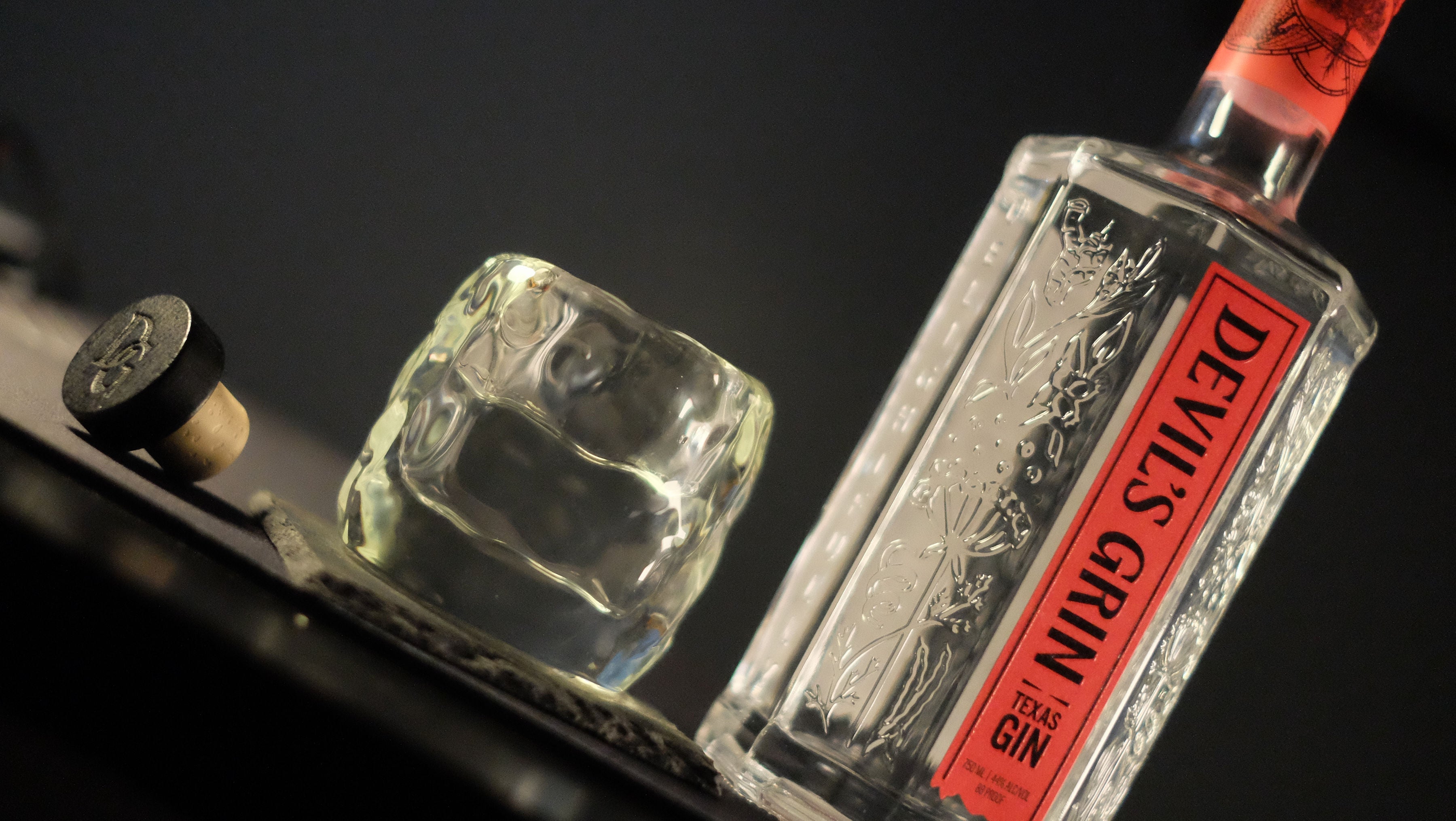 Close-up of Devil's Grin Texas Gin bottle with embossed botanical glass design alongside a rocks glass and cork stopper on a dark surface