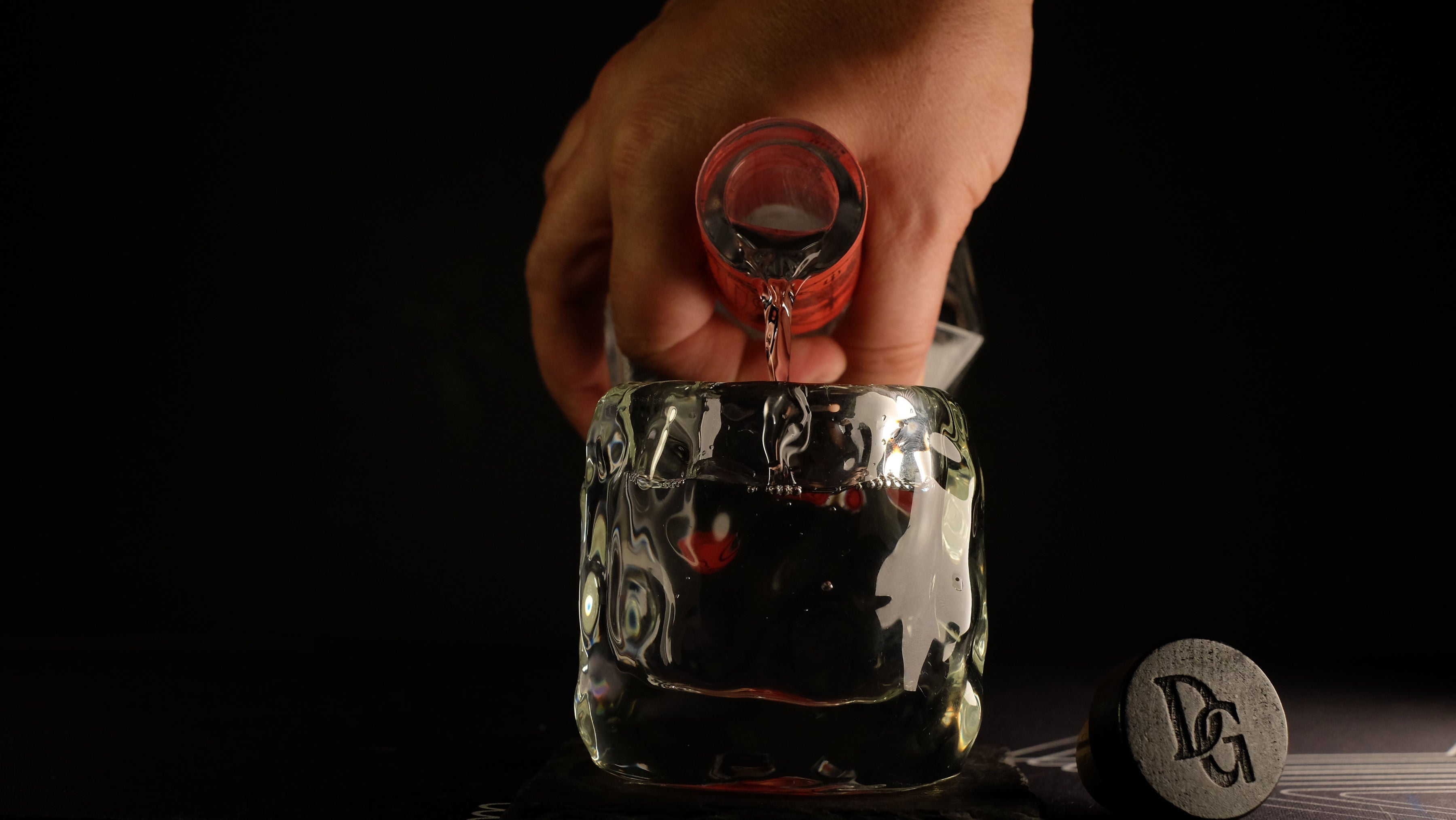 Hand pouring Devil's Grin Texas Gin over a large ice cube in a rocks glass with DG-branded cork on a dark background