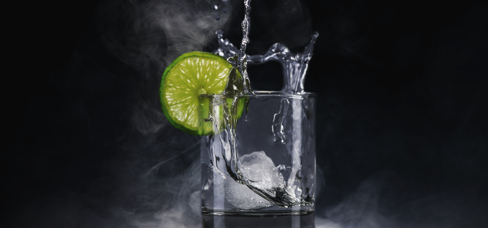Clear glass with ice, lime slice, and splashing liquid, creating a misty effect.