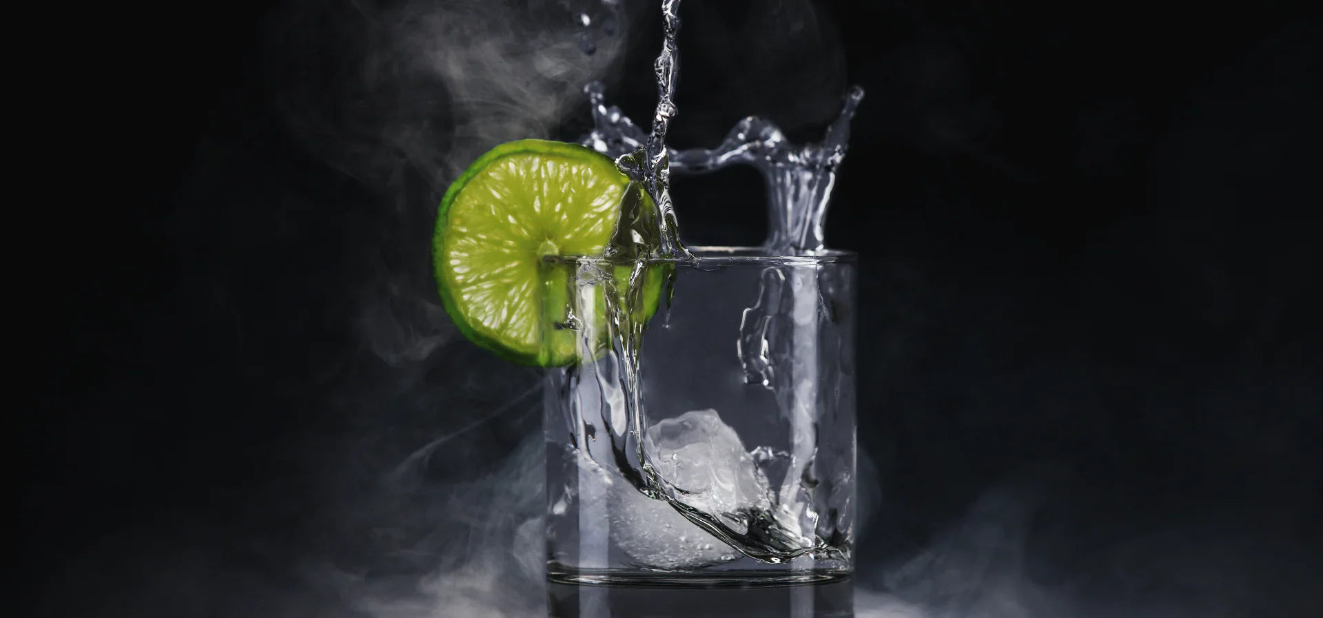 Clear glass with ice, lime slice, and splashing liquid, creating a misty effect.