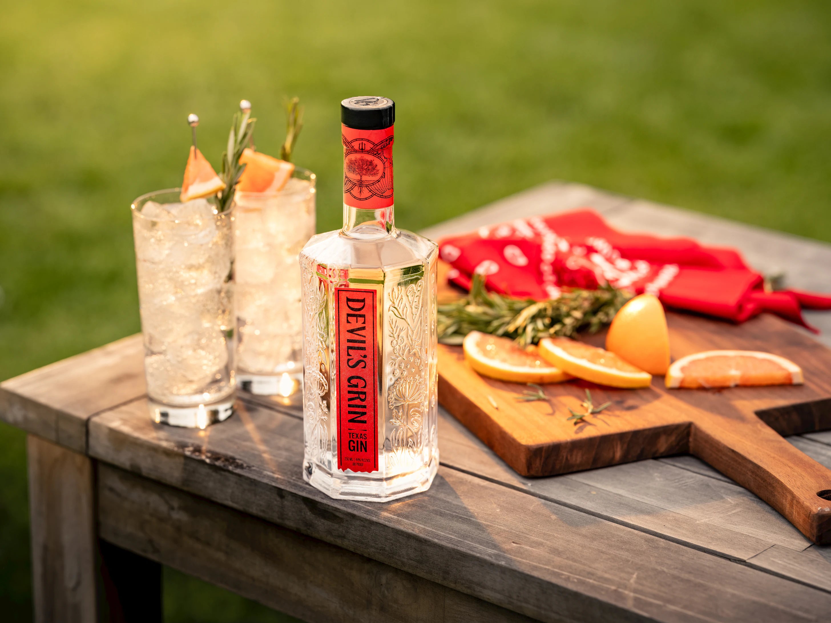 Devil's Grin Texas Gin bottle with gin and tonic cocktails, citrus slices, and rosemary on an outdoor wooden table