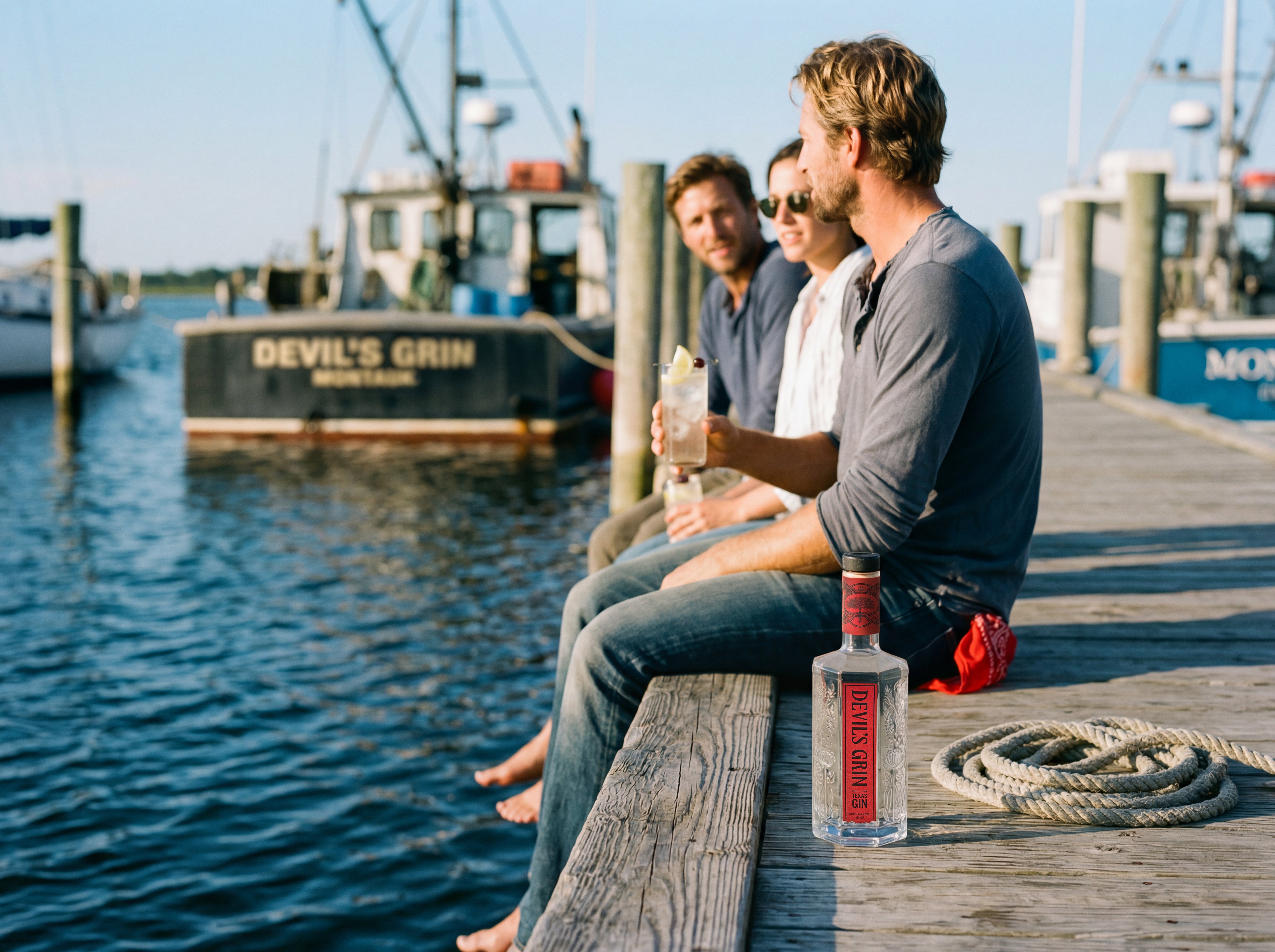 Friends enjoying Devil's Grin Texas Gin Tom Collins cocktails on a sunny harbor dock with fishing boat in background