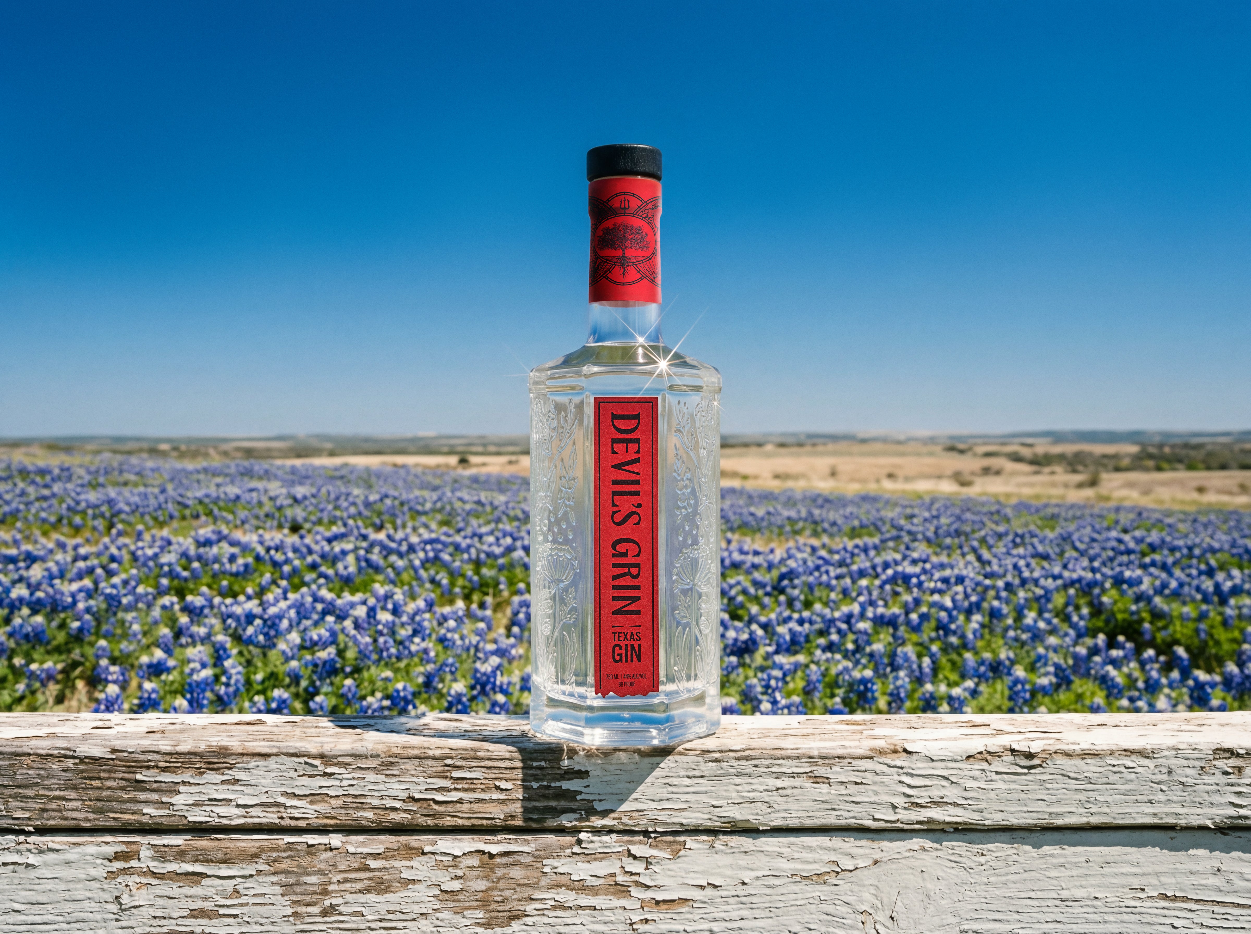 Devil's Grin Texas Gin bottle on rustic fence rail against blooming bluebonnet field and clear blue sky