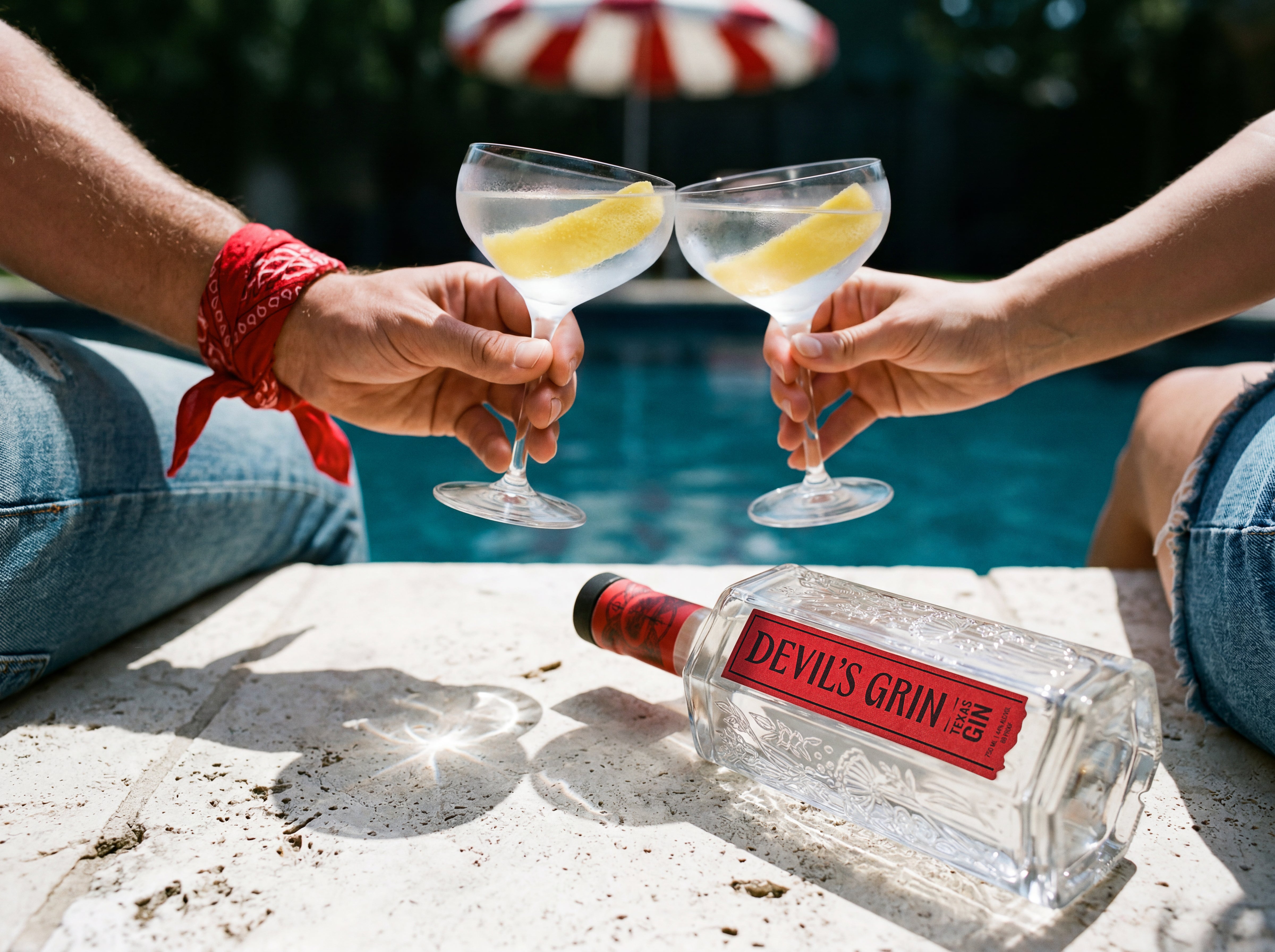 Two people toasting with lemon-garnished gin martinis poolside with a bottle of Devil's Grin Texas Gin