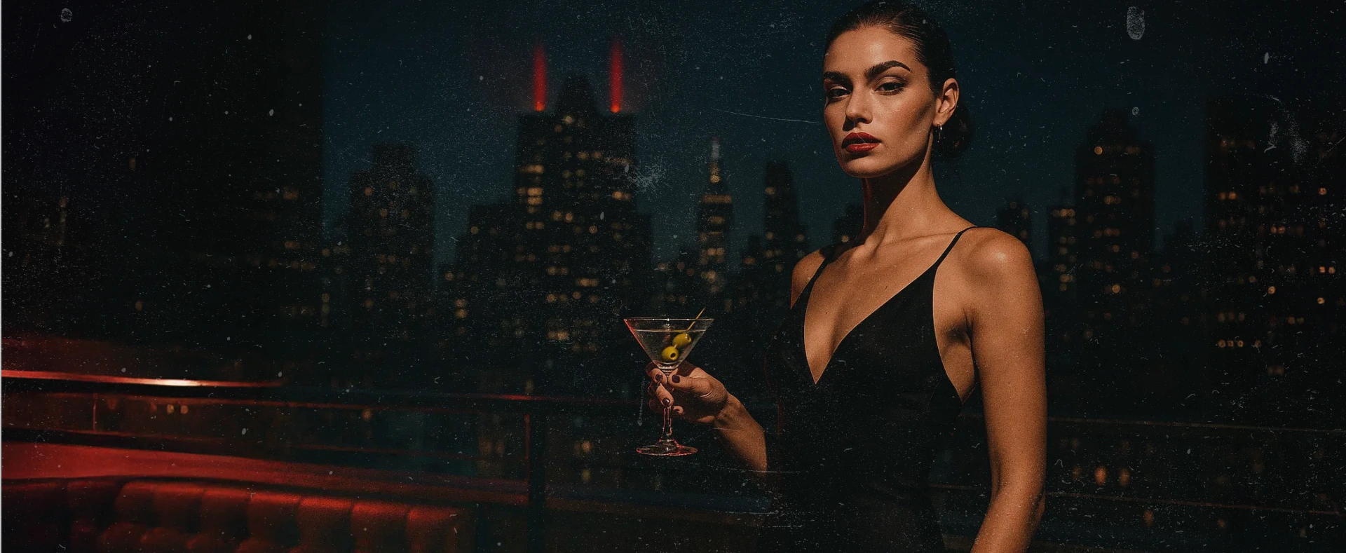 Glamorous woman in a black dress holds a martini on a city rooftop at night with red lighting and a city skyline